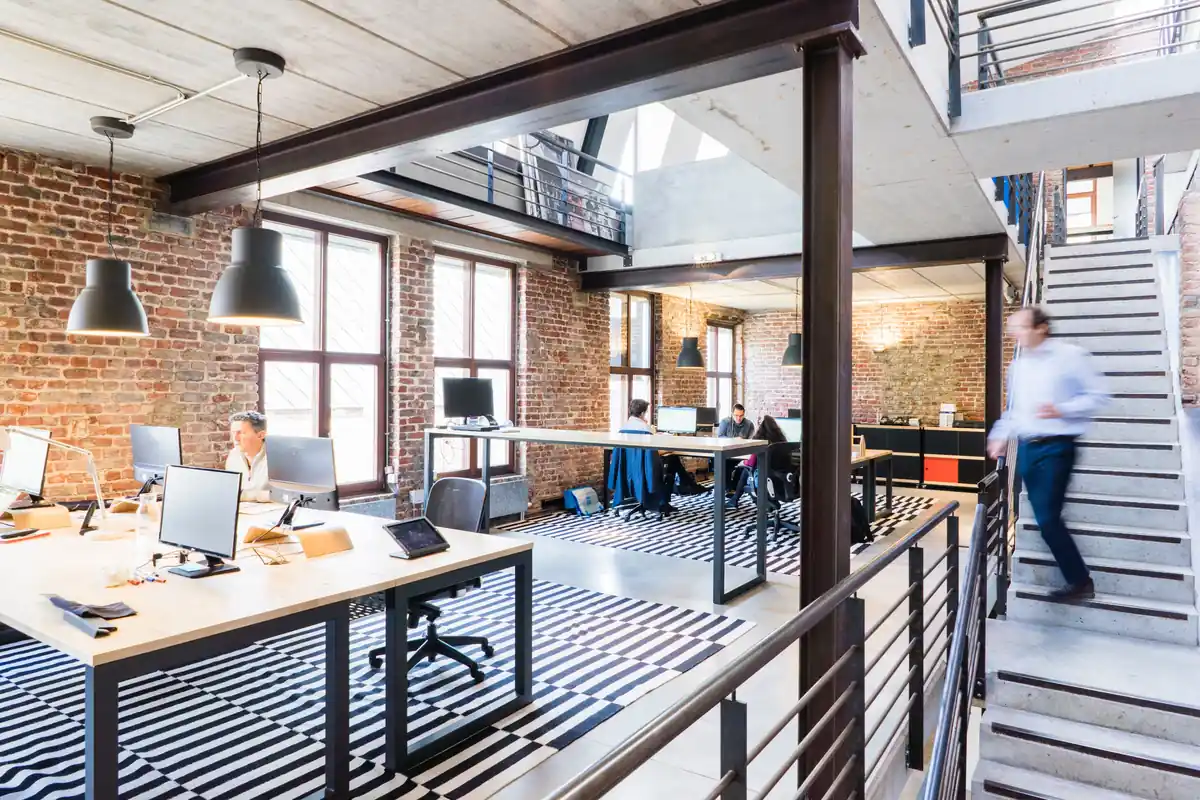 Open loft coworking floor with exposed brick, long shared desks, and a staircase