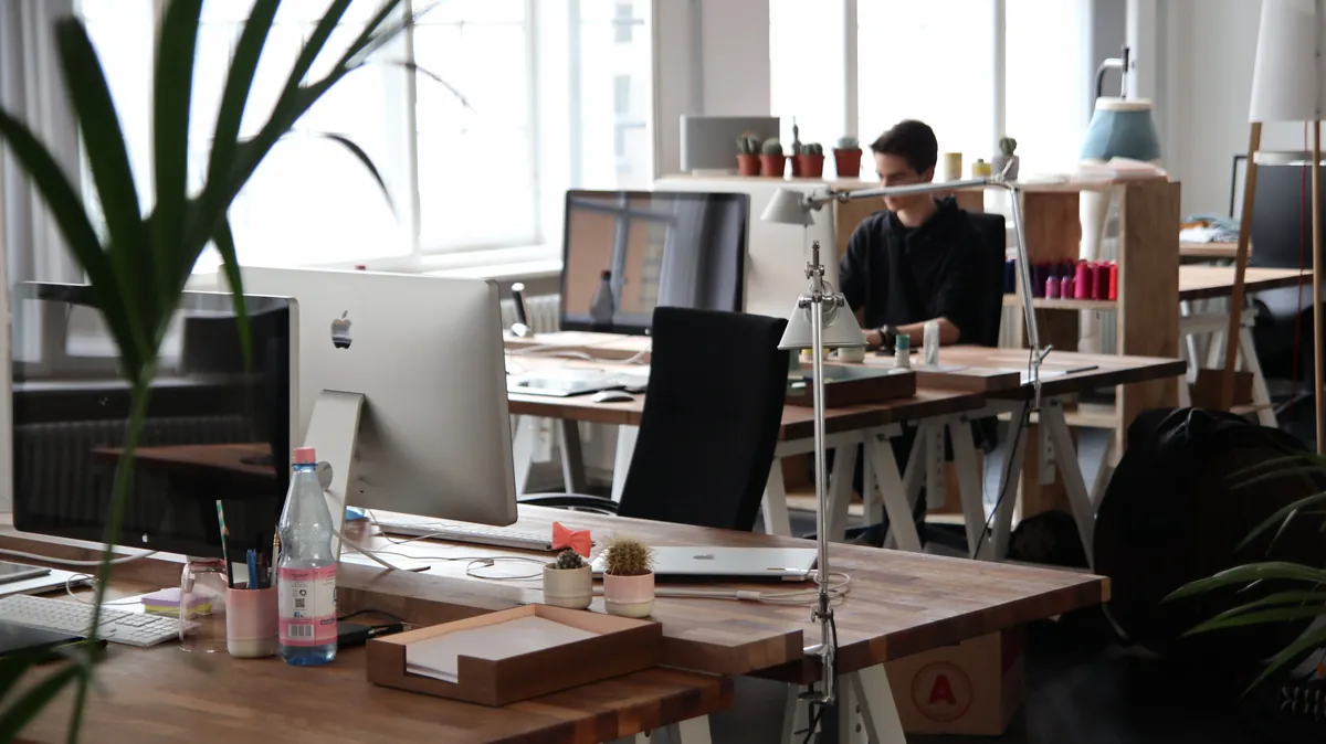 Shared workspace floor with wooden trestle desks, computers, and daylight from tall windows